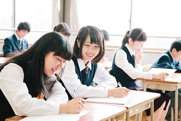 High school students, seven teenagers study in a classroom, Japan. Interior shot, sitting and writing at a desk, young people, in a row, horizontal composition. Focus on the foreground.